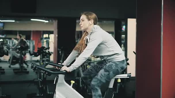 Krásná mladá dívka v tělocvičně, třást nohy na cyklistický trenažér, usmívají se na kameru. Koncept: milovat sport, navštěvovat tělocvičnu, správná výživa, štíhlé tělo, aby se zdravá. 4k — Stock video