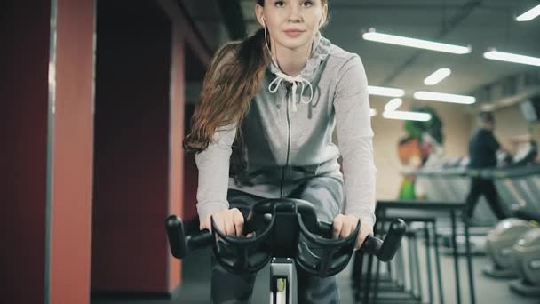 Молодая красивая девушка в спортзале, трясет ногами на велосипедном тренажере, улыбается в камеру. Концепция: любить спорт, посещать тренажерный зал, правильное питание, стройное тело, быть здоровым. 4k — стоковое видео