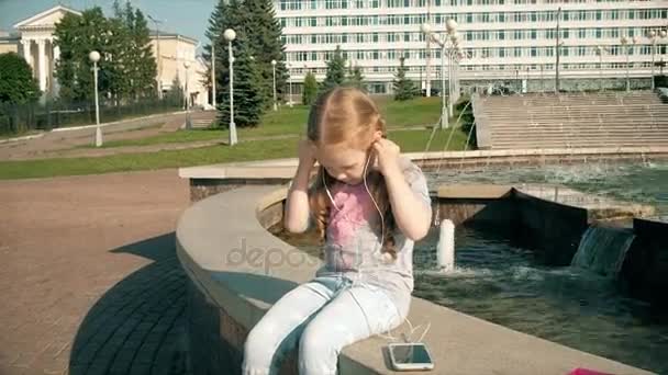 Hermosa chica escuchando música en los auriculares con teléfono cerca de la fuente — Vídeos de Stock