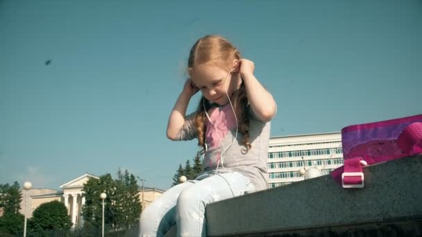 Beautiful girl listening to music on headphones with phone near fountain — Stock Video
