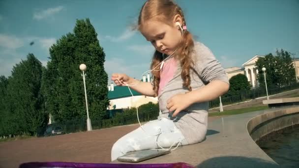 Krásná dívka poslouchat hudbu z sluchátka s telefonem poblíž fountain — Stock video