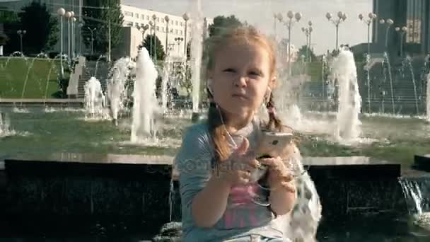 美しい少女の噴水の近くの携帯電話とヘッドフォンで音楽を聴く — ストック動画