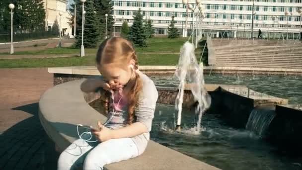 Mooi meisje, luisteren naar muziek op de hoofdtelefoon met de telefoon in de buurt van fontein — Stockvideo