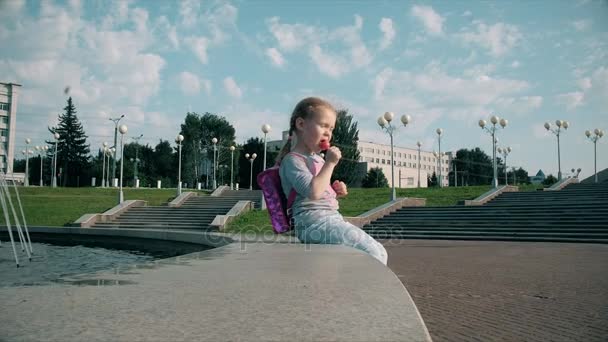 Gyönyörű lány eszik nyalóka a szökőkút közelében a fényes nap — Stock videók