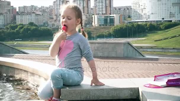 Mooi meisje lollipop eten in de buurt van een fontein op een heldere dag — Stockvideo