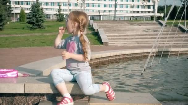 Beautiful girl eating lollipop near a fountain on a bright day — Stock Video