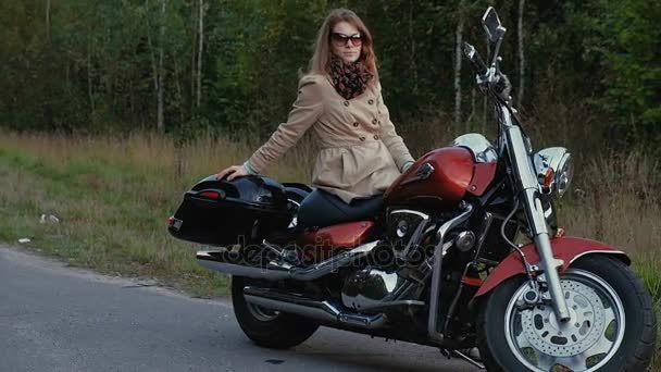 Ung tjej med brunt hår sitter på en motorcykel nära en väg. — Stockvideo