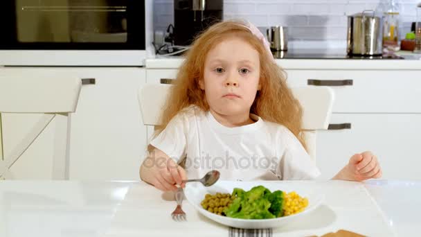 Une petite belle fille mange du brocoli et des petits pois verts avec plaisir, à table à la maison — Video