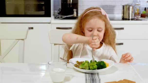 En liten vacker flicka äter broccoli och gröna ärtor med nöje, vid bordet hemma — Stockvideo