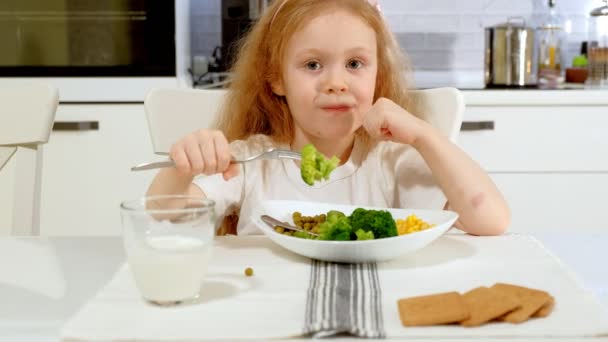 Маленькая красивая девушка ест брокколи и зеленый горох с удовольствием, за столом у себя дома — стоковое видео
