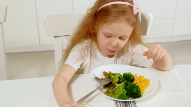 Uma menina bonita pequena come brócolis e ervilhas verdes com prazer, na mesa em casa — Vídeo de Stock