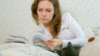 Bir kadın yatakta bir kitap okuyor, Gri kedi onun yanında yatıyor