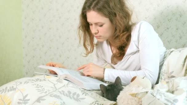 Una donna sta leggendo un libro a letto, un gatto grigio è sdraiato accanto a lei — Video Stock