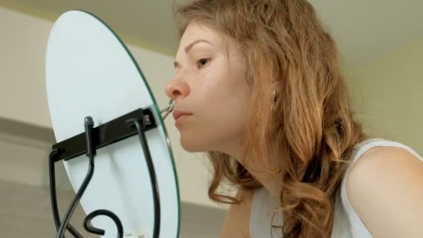 Молодая женщина надевает тушь и смотрит в ручное зеркало в спальне дома — стоковое видео