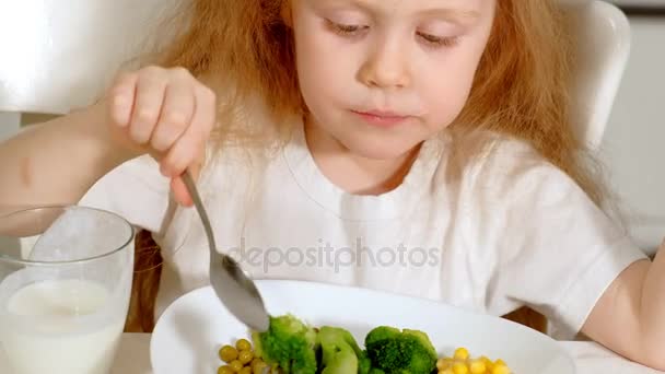 Una pequeña chica hermosa come brócoli y guisantes verdes con placer, en la mesa en casa — Vídeos de Stock