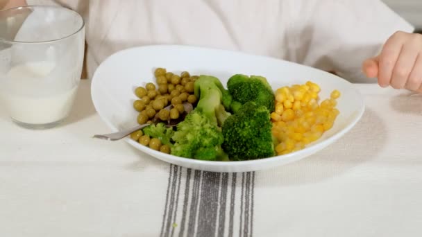 Una piccola bella ragazza mangia con piacere broccoli e piselli verdi, a tavola a casa — Video Stock