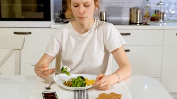 Una hermosa mujer come brócoli y guisantes verdes con placer, en la mesa en casa — Vídeos de Stock