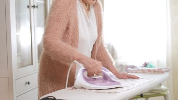 Старая женщина гладит одежду дома — стоковое видео
