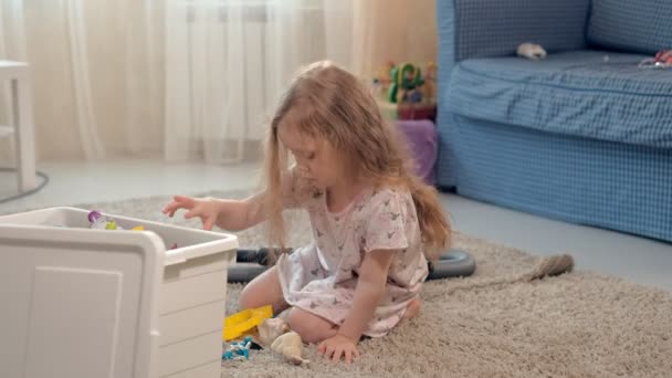 Девушка собирает игрушки в коробке, сидя дома на полу — стоковое видео
