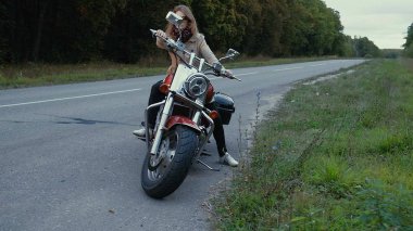 Kahverengi saçlı genç kız bir road yakınındaki bir motosiklet üzerinde oturur.