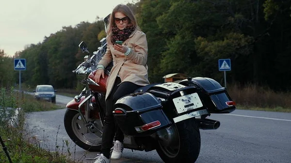 Chica joven con el pelo castaño se sienta en una moto cerca de una carretera . — Foto de Stock