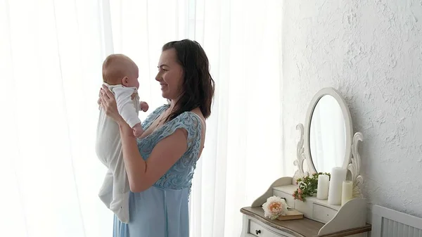 Seguindo a mãe colocando o bebê para dormir enquanto caminha. Mulher atraente segurando bebê em mãos e andar através da sala de estar com cortinas de janela brilhantes no fundo . — Fotografia de Stock
