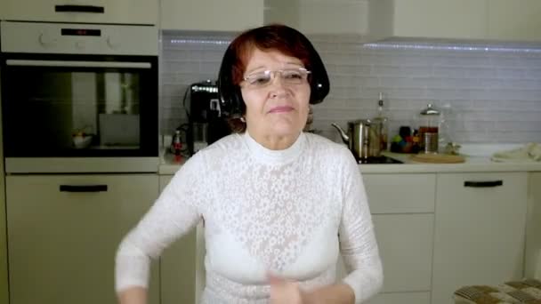 An elderly woman listens to music on headphones from the phone, gesticulates energetically and dances — Stock Video