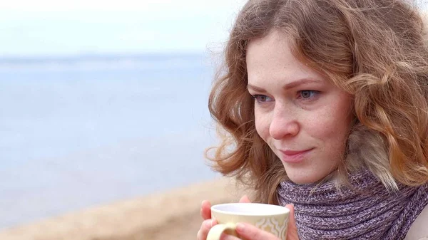 Romantische junge Frau, die es sich am Strand gemütlich macht, heißen Tee oder Kaffee aus der Thermoskanne trinkt. ruhiger und gemütlicher Abend. — Stockfoto