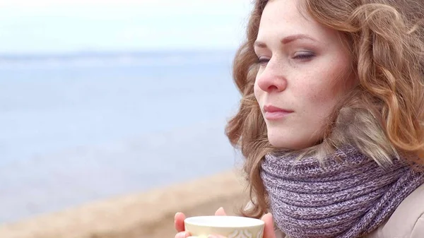 Romantische junge Frau, die es sich am Strand gemütlich macht, heißen Tee oder Kaffee aus der Thermoskanne trinkt. ruhiger und gemütlicher Abend. — Stockfoto