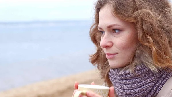 Romantische junge Frau, die es sich am Strand gemütlich macht, heißen Tee oder Kaffee aus der Thermoskanne trinkt. ruhiger und gemütlicher Abend. — Stockfoto