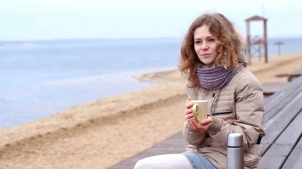 Romantische junge Frau, die es sich am Strand gemütlich macht, heißen Tee oder Kaffee aus der Thermoskanne trinkt. ruhiger und gemütlicher Abend. — Stockfoto