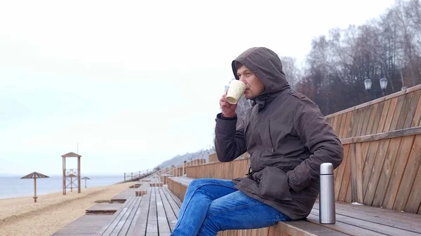 Romantico giovane uomo che si rilassa sulla spiaggia con, bere tè caldo o caffè dal thermos. Serata tranquilla e accogliente . — Foto Stock