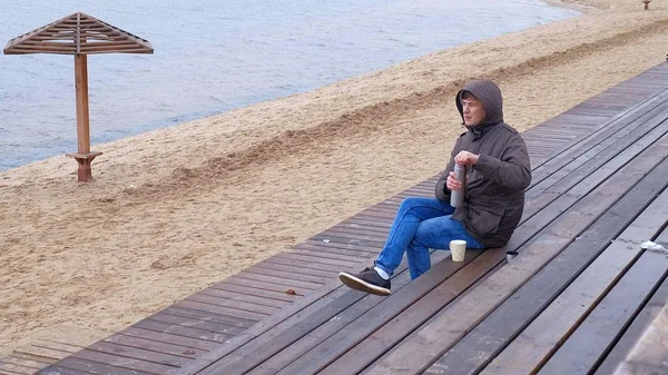 Romantico giovane uomo che si rilassa sulla spiaggia con, bere tè caldo o caffè dal thermos. Serata tranquilla e accogliente . — Foto Stock