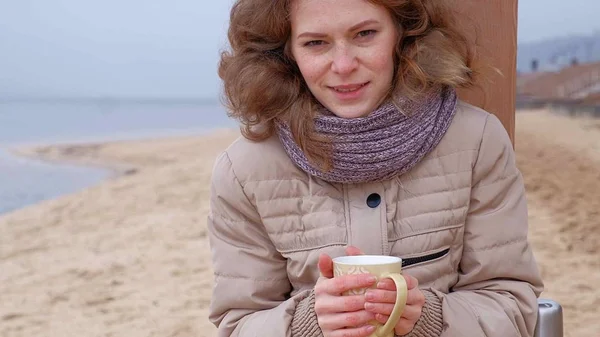 Romantische junge Frau, die es sich am Strand gemütlich macht, heißen Tee oder Kaffee aus der Thermoskanne trinkt. ruhiger und gemütlicher Abend. — Stockfoto