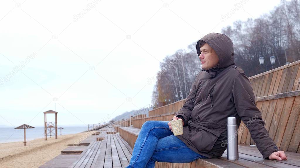 Romantic young man relaxing on the beach with , drinking hot tea or coffee from thermos. Calm and cozy evening.