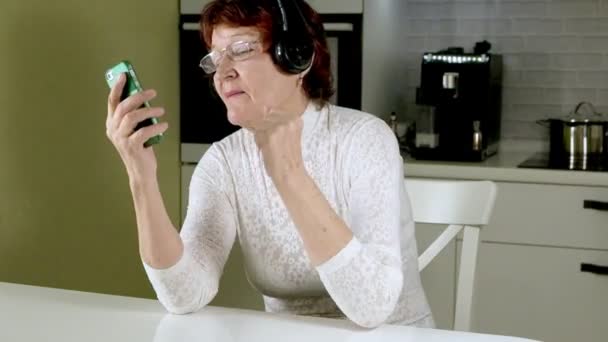 Een oudere vrouw luistert naar muziek op hoofdtelefoon uit de telefoon, gesticulates, energiek en danst — Stockvideo