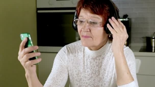 Eine ältere Frau hört über Kopfhörer Musik aus dem Telefon, gestikuliert energisch und tanzt — Stockvideo