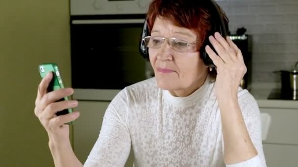 An elderly woman listens to music on headphones from the phone, gesticulates energetically and dances — Stock Video