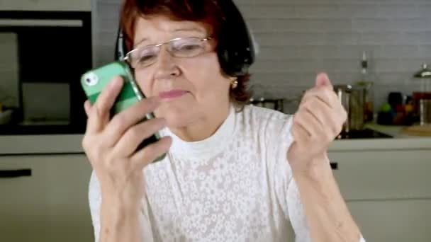 An elderly woman listens to music on headphones from the phone, gesticulates energetically and dances — Stock Video