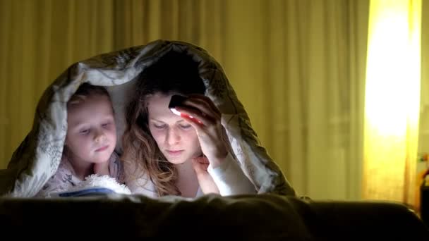 Glimlachend moeder en haar dochter lezen boek onder bed en houden van een zaklamp. — Stockvideo