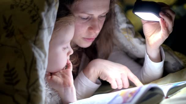 Sonriente madre y su hija leyendo libro bajo la cubierta de la cama y sosteniendo una linterna . — Vídeo de stock