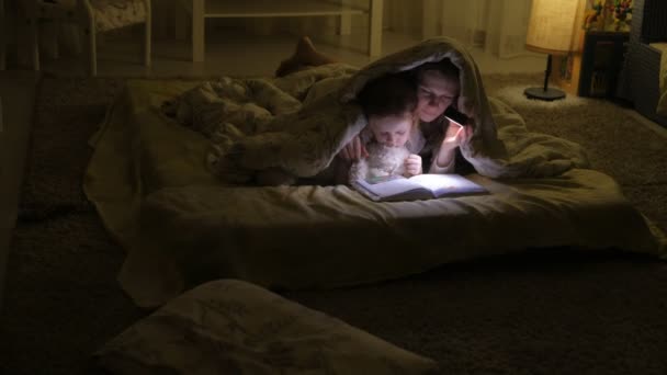 Sorrindo mãe e sua filha lendo livro sob a capa da cama e segurando uma lanterna . — Vídeo de Stock