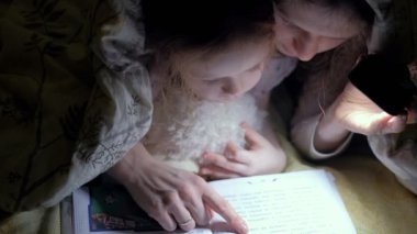 Anne ve kızı yatak örtüsü altında kitap okuma ve bir el feneri tutan gülümseyerek.