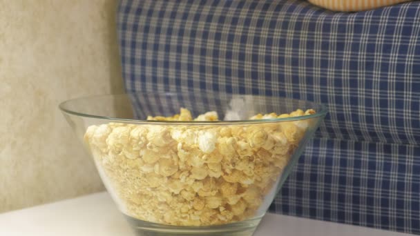 Una niña está sentada en el sofá viendo la televisión y comiendo palomitas de maíz. — Vídeos de Stock