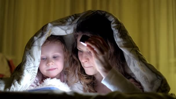Улыбающаяся мать и ее дочь читают книгу под одеялом и держат фонарик . — стоковое видео