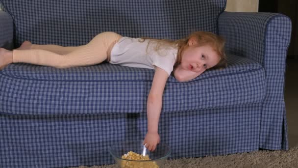 Una niña está sentada en el sofá viendo la televisión y comiendo palomitas de maíz. — Vídeo de stock