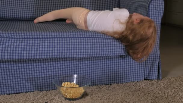 Una niña está sentada en el sofá viendo la televisión y comiendo palomitas de maíz. — Vídeos de Stock