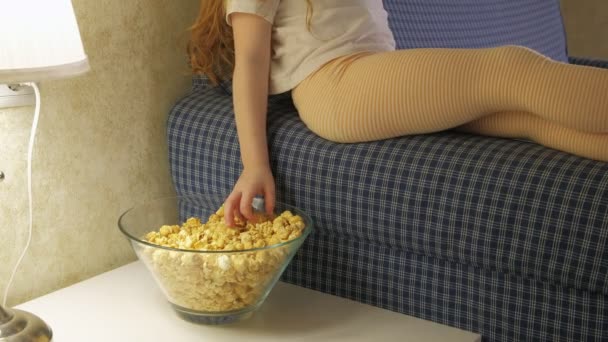 Una niña está sentada en el sofá viendo la televisión y comiendo palomitas de maíz. — Vídeos de Stock