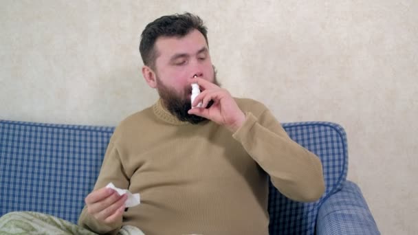 A man with a cold sits on the couch. He sprinkles a special nasal spray into his nose. — Stock Video