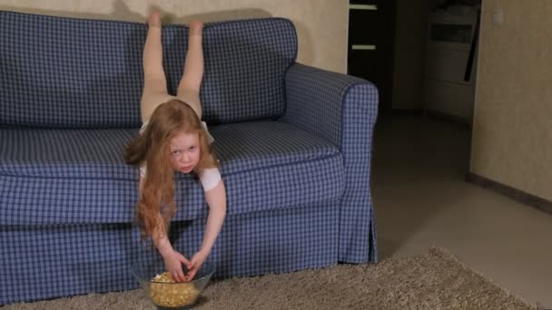 Una niña está sentada en el sofá viendo la televisión y comiendo palomitas de maíz. — Vídeos de Stock